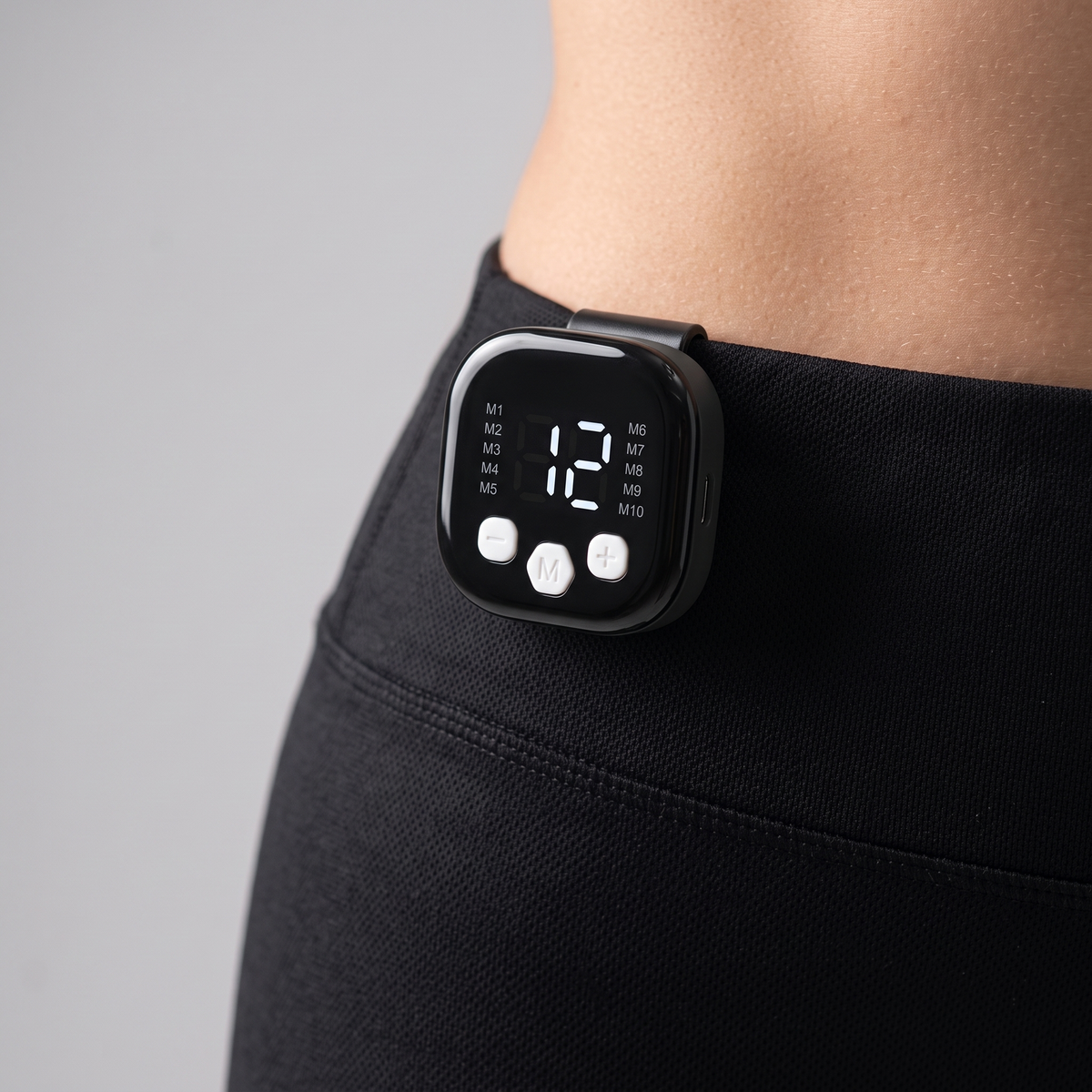 Boîtier de commande du short EMS clipsé sur la ceinture : écran LED avec mode M1–M10 et niveau d'intensité, touches +, M et −